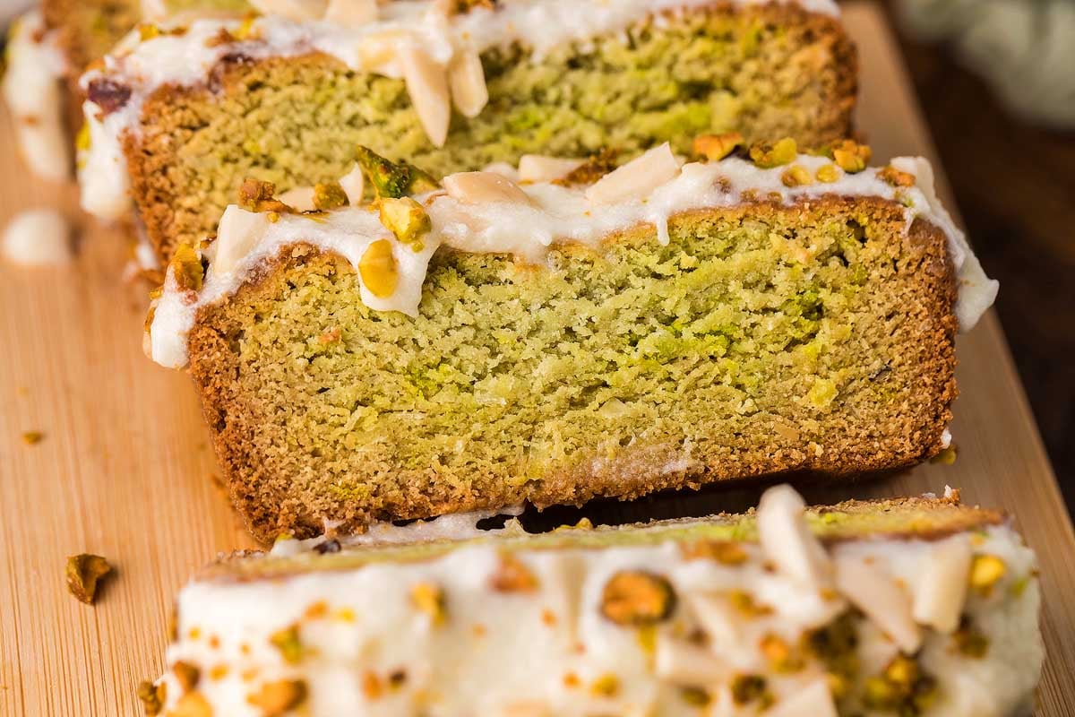 Close-up of sliced pistachio loaf cake with white icing and chopped nuts on a wooden board.