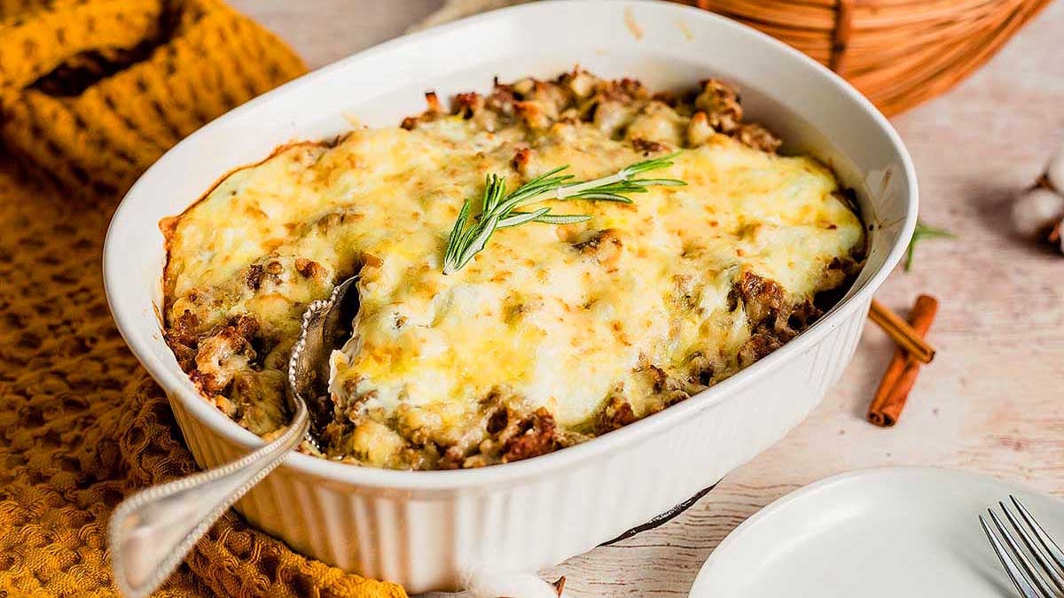 A baked casserole topped with melted cheese and a rosemary sprig, with a serving spoon in the dish.