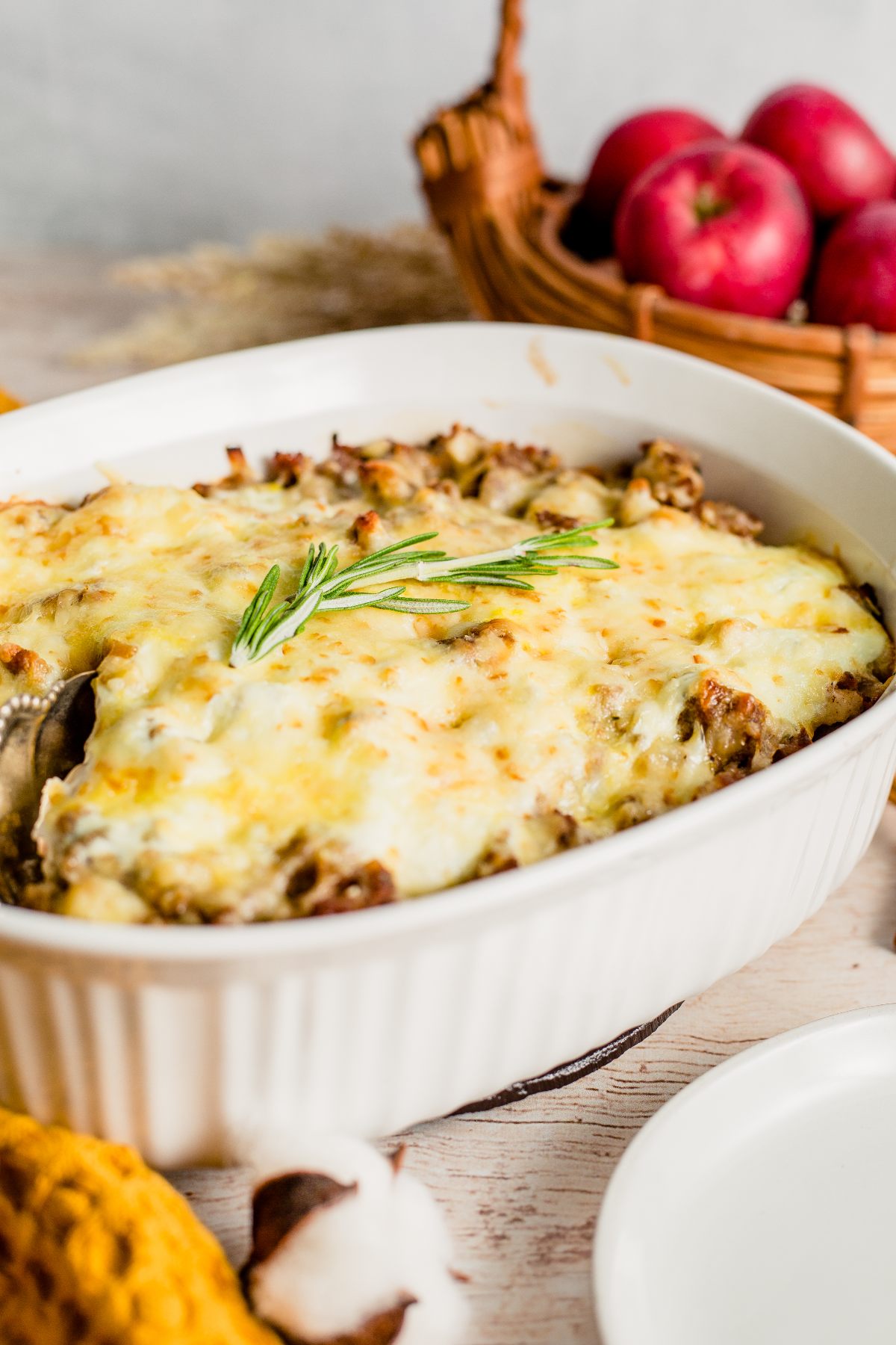 A baked breakfast casserole with apples, topped with melted cheese and garnished with rosemary, with apples in a basket in the background. An easy family brunch recipe everyone will love.