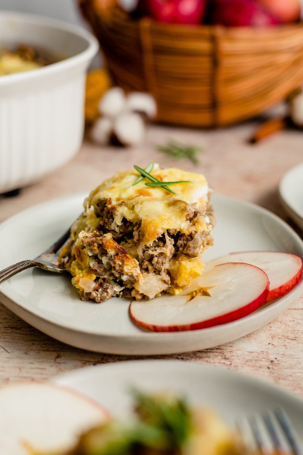 A slice of cheesy Sausage Apple Breakfast Casserole on a plate with apple slices, garnished with herbs, and a fork beside it—an easy family brunch recipe everyone will love.