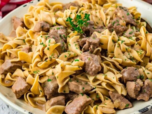 A plate of creamy slow cooker beef noodle casserole with egg noodles and beef chunks, garnished with chopped parsley. A red and white cloth is partially visible on the side.