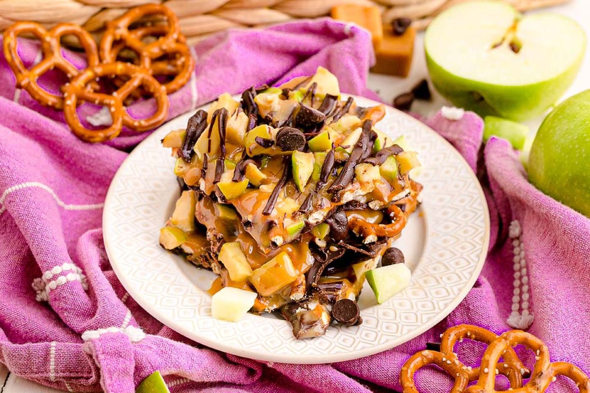 A plate of caramel apple pretzel bark sits on a white decorative plate, drizzled with chocolate and garnished with green apple chunks and chocolate chips. The plate rests on a purple cloth, surrounded by pretzels, green apple pieces, and caramel squares.