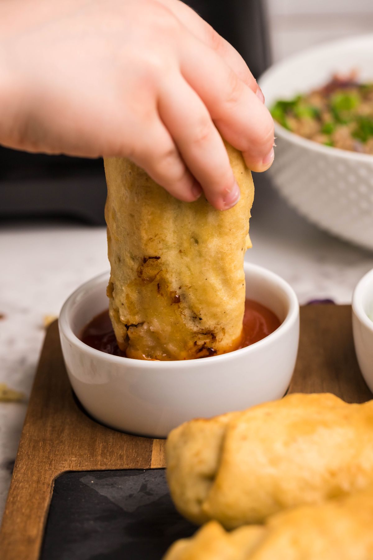 A hand dips a fried snack made with Air Fryer Gluten Free Egg Roll Wrappers into a small bowl of red sauce on a wooden tray.