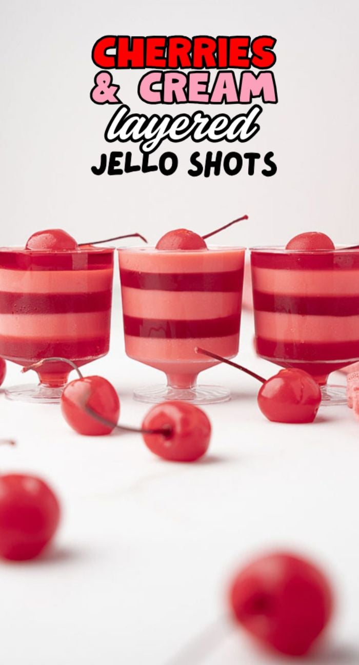 Layered rum jello shots with cherry rum and vanilla rum, topped with a cherry and scattered cherries around—these Cherries and Cream Jello Shots are perfect for any festive gathering.