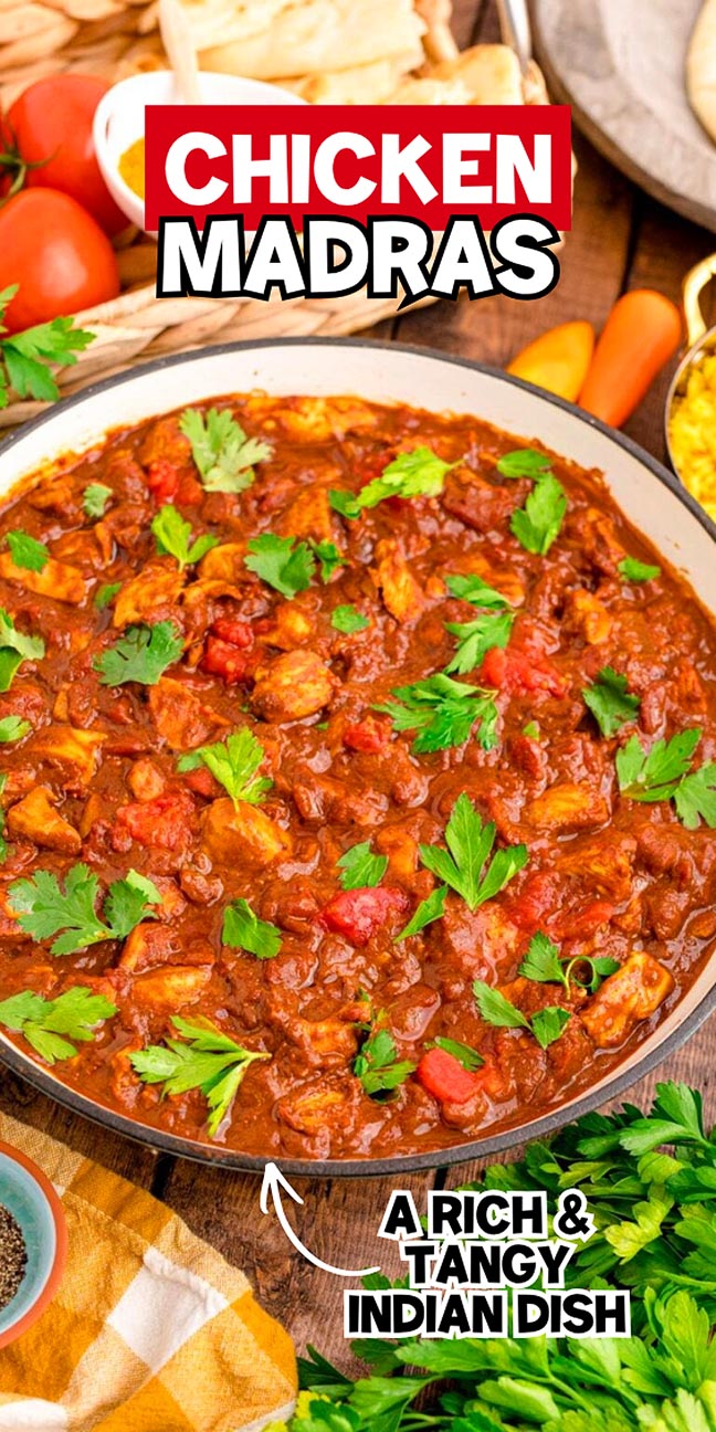 A bowl of chicken madras garnished with parsley, surrounded by tomatoes and spices on a wooden table.