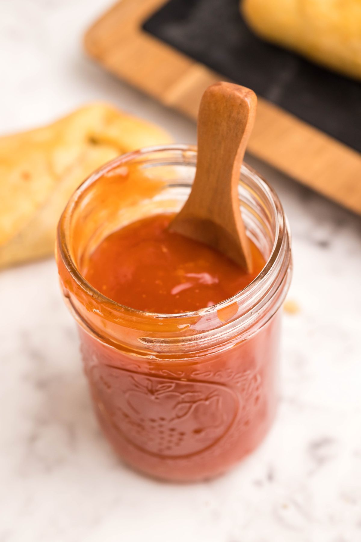 A glass jar of orange-red sweet and sour sauce with a wooden spoon inside, sitting on a marble surface.