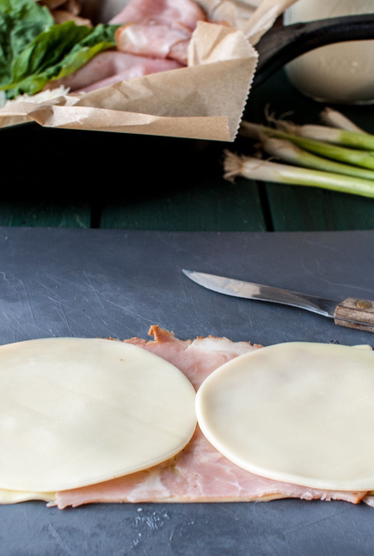 Two slices of cheese on top of deli ham on a cutting board, with a knife and green onions nearby—perfect for making Ham and Provolone Rotolo – Lasagna Roll Ups.