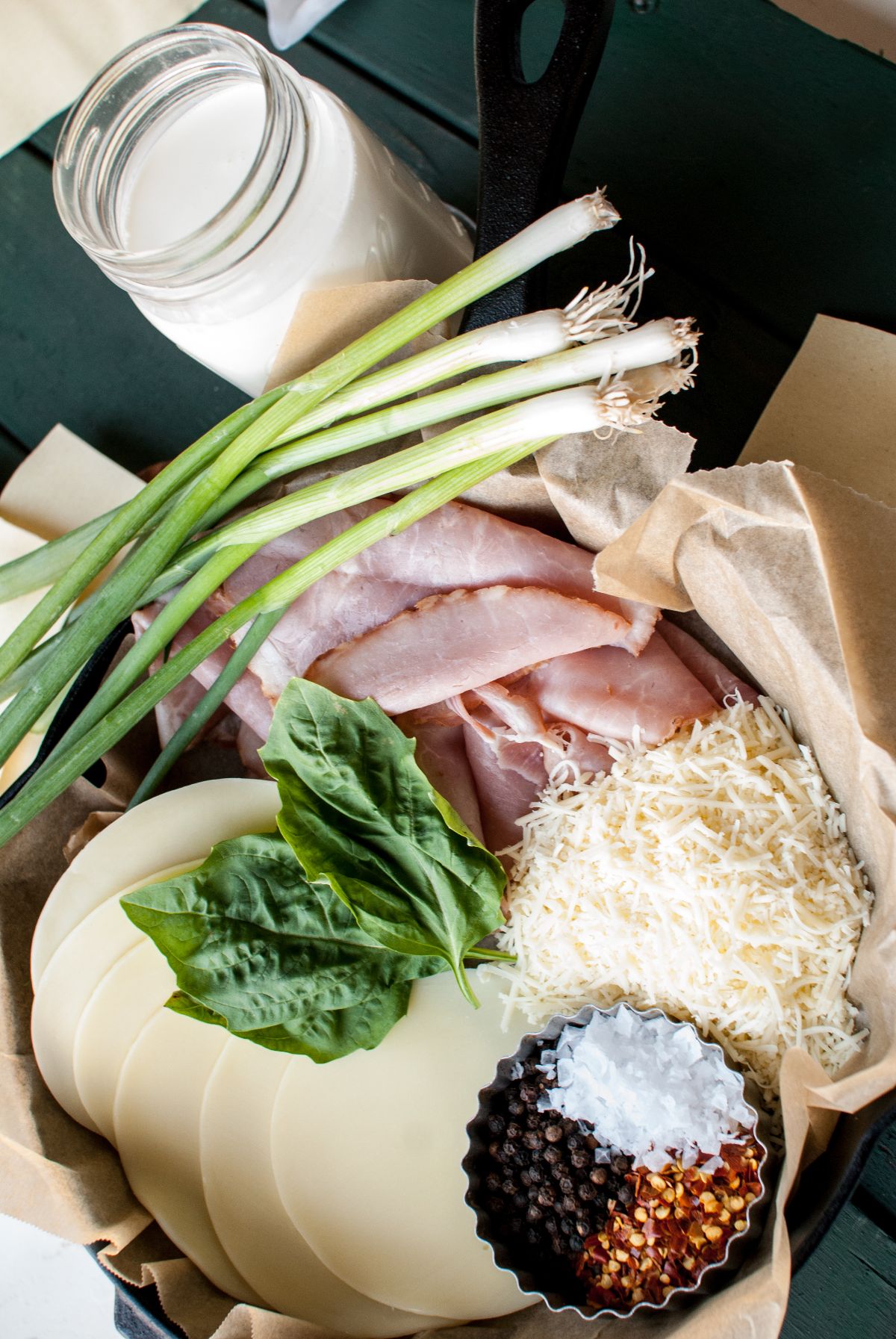 Sliced ham, cheese, shredded cheese, green onions, basil, spices, and a jar of milk on parchment paper—perfect ingredients for Baked pasta roses or Ham and Provolone Rotolo – Lasagna Roll Ups.