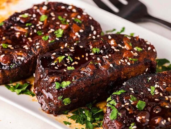Three slow cooker pork chops with a sweet and sticky Chinese-inspired glaze, garnished with chopped herbs and sesame seeds, served on a white rectangular plate.