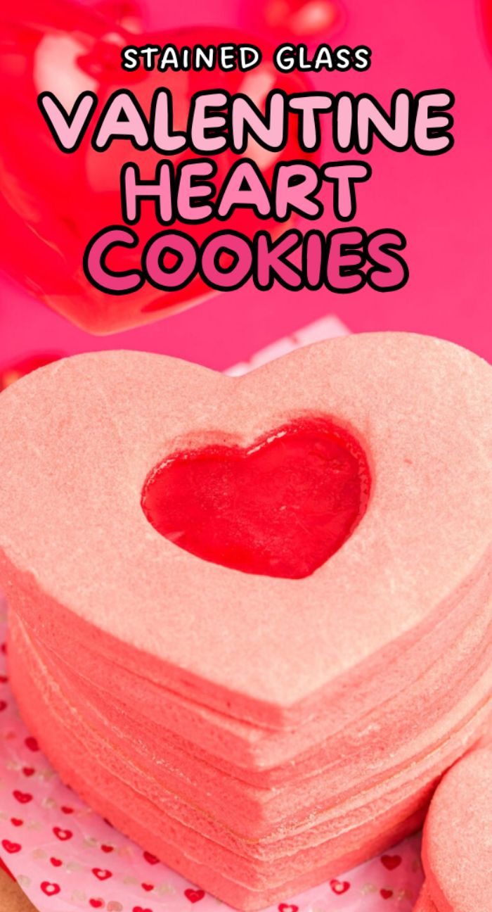 Stack of heart shaped sugar cookies with a translucent red center made with Jolly Ranchers, on a pink surface. Text above reads "Stained Glass Valentine Heart Cookies.