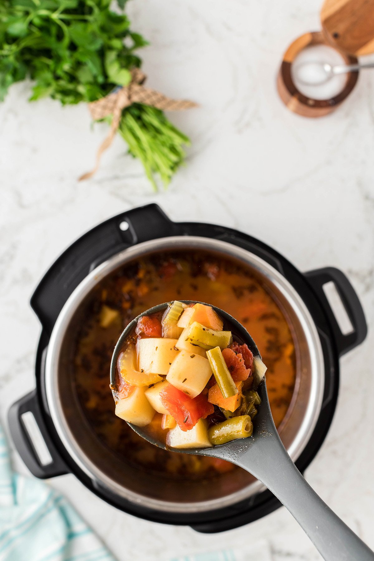 A ladle of vegetable soup above an Instant Pot, made with fresh and frozen vegetables, with fresh herbs and salt nearby on a marble countertop.