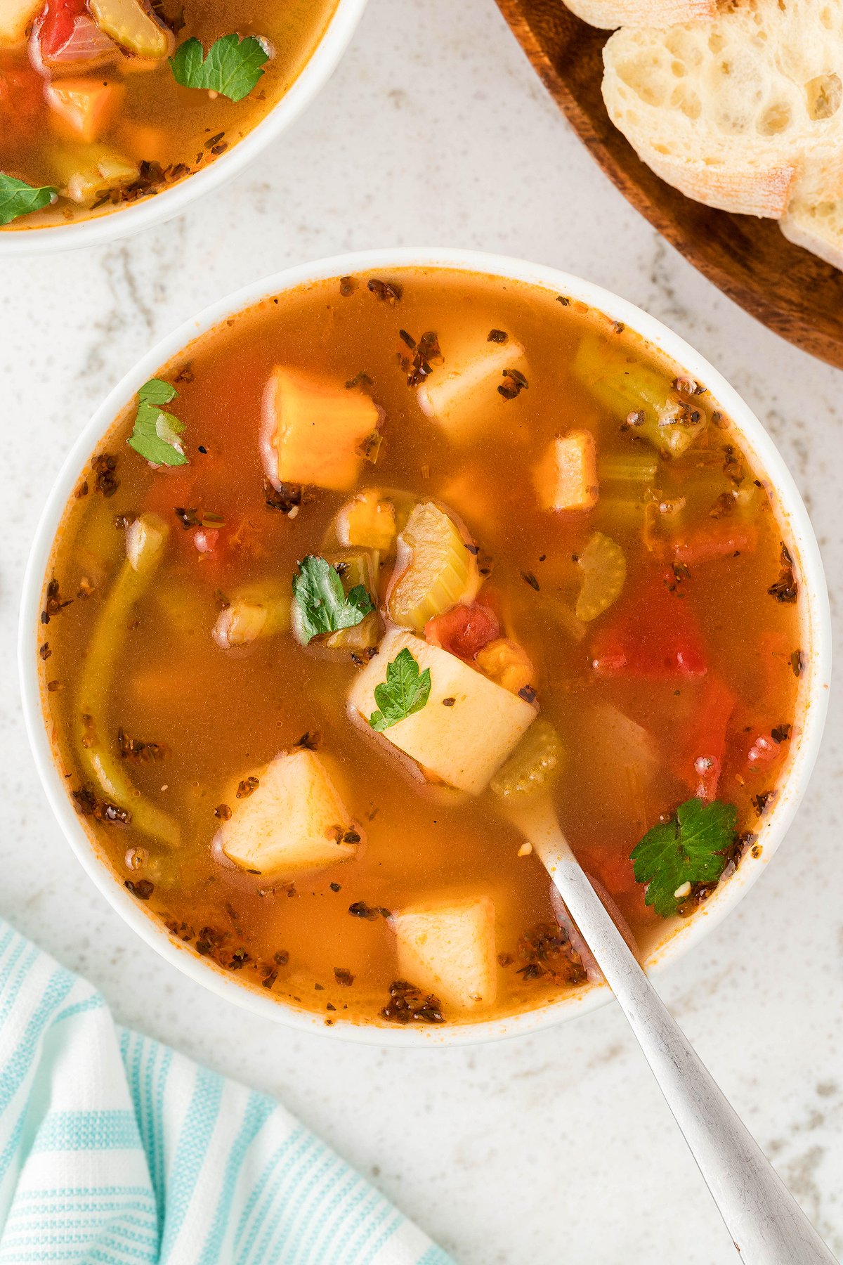 Bowl of Instant Pot Vegetable Soup with potatoes, celery, and parsley, made with fresh and frozen vegetables, next to sliced bread on a white surface.