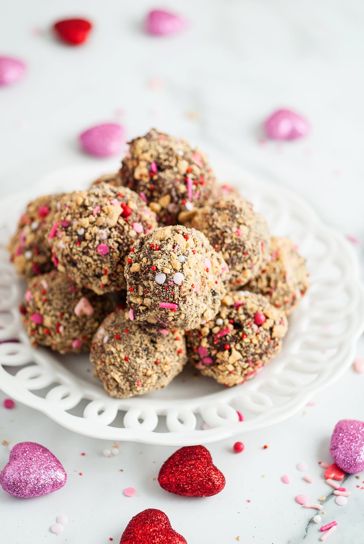 A plate of chocolate S’more truffles coated with nuts and colorful sprinkles, surrounded by glittery heart decorations—an easy Valentine treat everyone will love.
