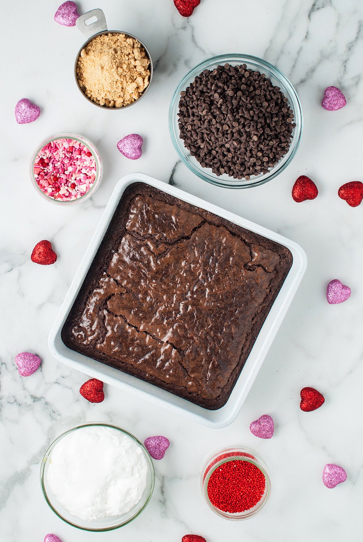 A pan of brownies surrounded by bowls of chocolate chips, brown sugar, sprinkles, and heart-shaped candies—perfect ingredients for making easy Valentine treat like Valentine S'mores Brownie Bites.