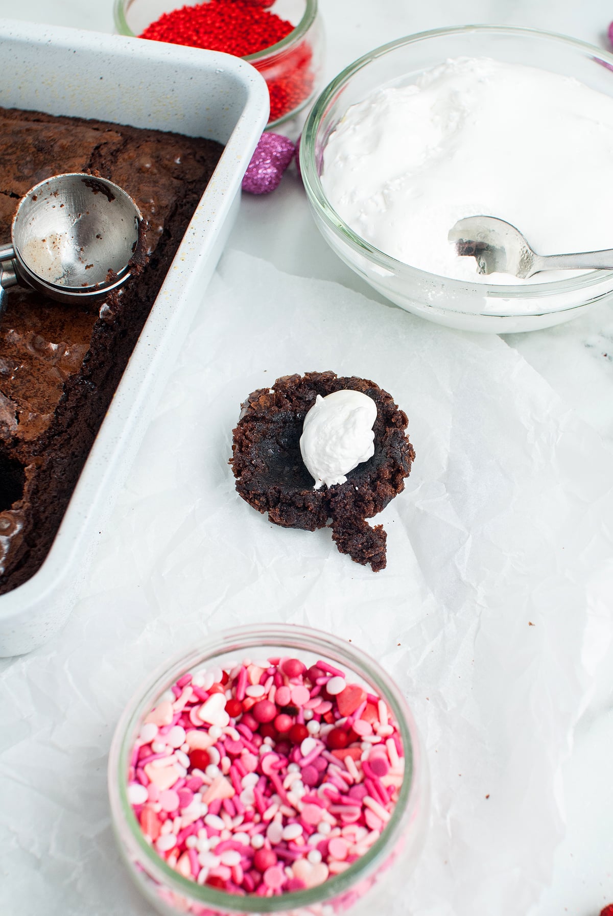 A brownie circle topped with white frosting, surrounded by sprinkles, brownies, and frosting bowls—these Valentine S'mores Brownie Bites are an easy Valentine treat, perfect for sharing and simple to make using a boxed brownie mix.