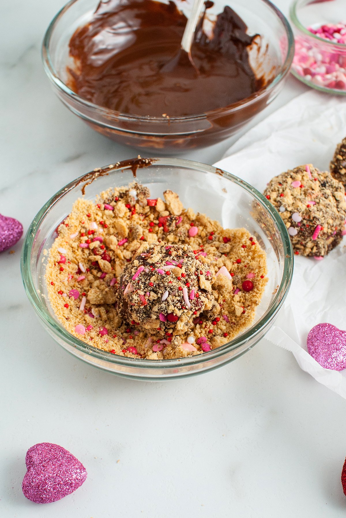 A chocolate truffle is being coated in crushed cookies and pink sprinkles, with melted chocolate in the background—an easy Valentine treat perfect for sharing.