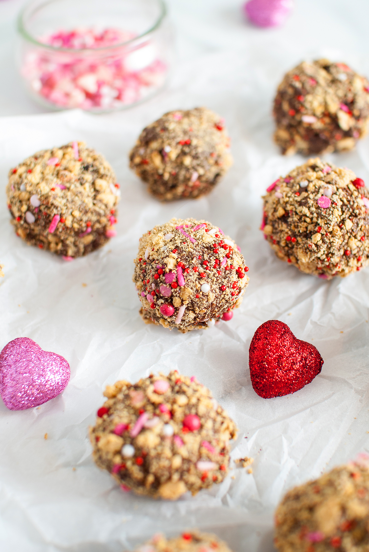 Chocolate truffles coated with sprinkles and crumbs, surrounded by glittery red and pink heart decorations—an easy Valentine treat inspired by S’more truffles.