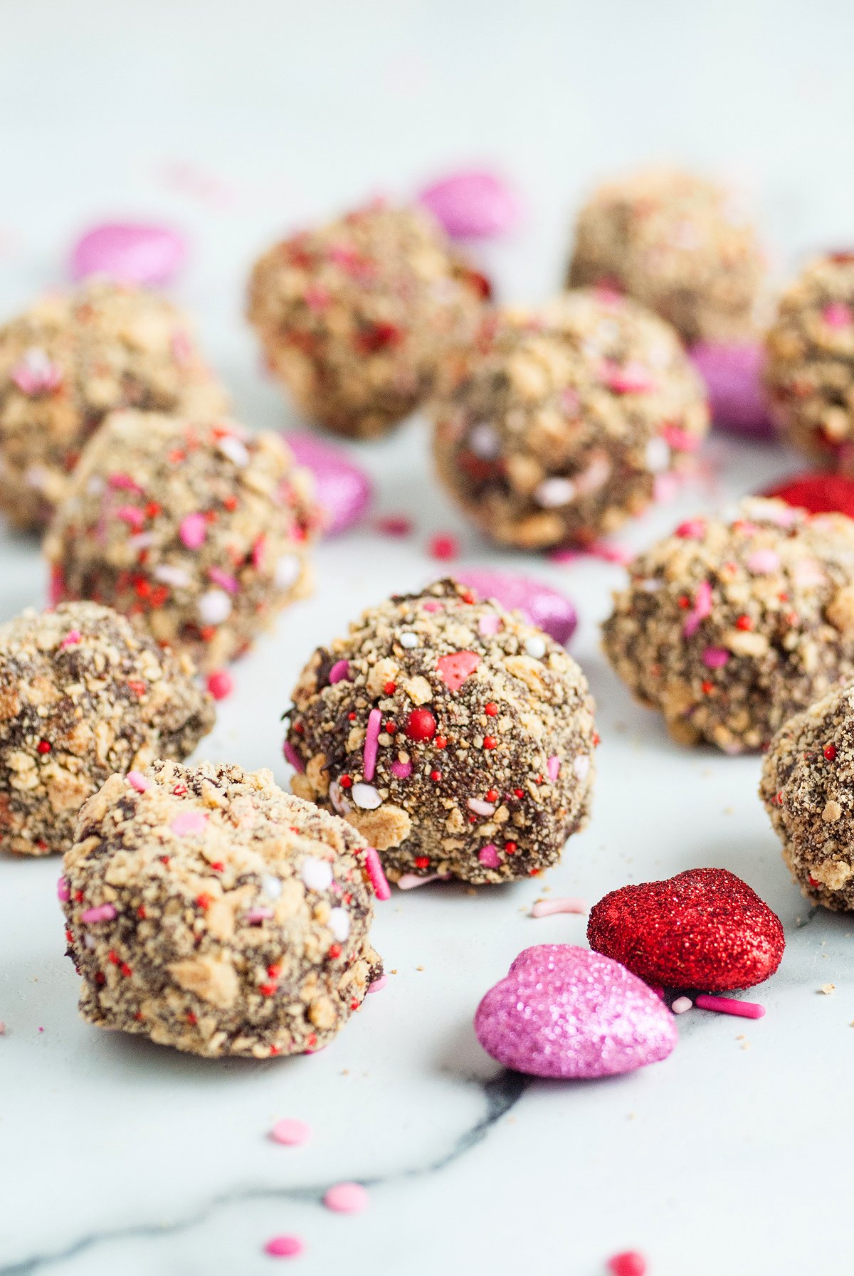 Chocolate truffles coated in sprinkles, with pink and red candy hearts scattered on a white surface—an easy Valentine treat, especially when made using a boxed brownie mix.