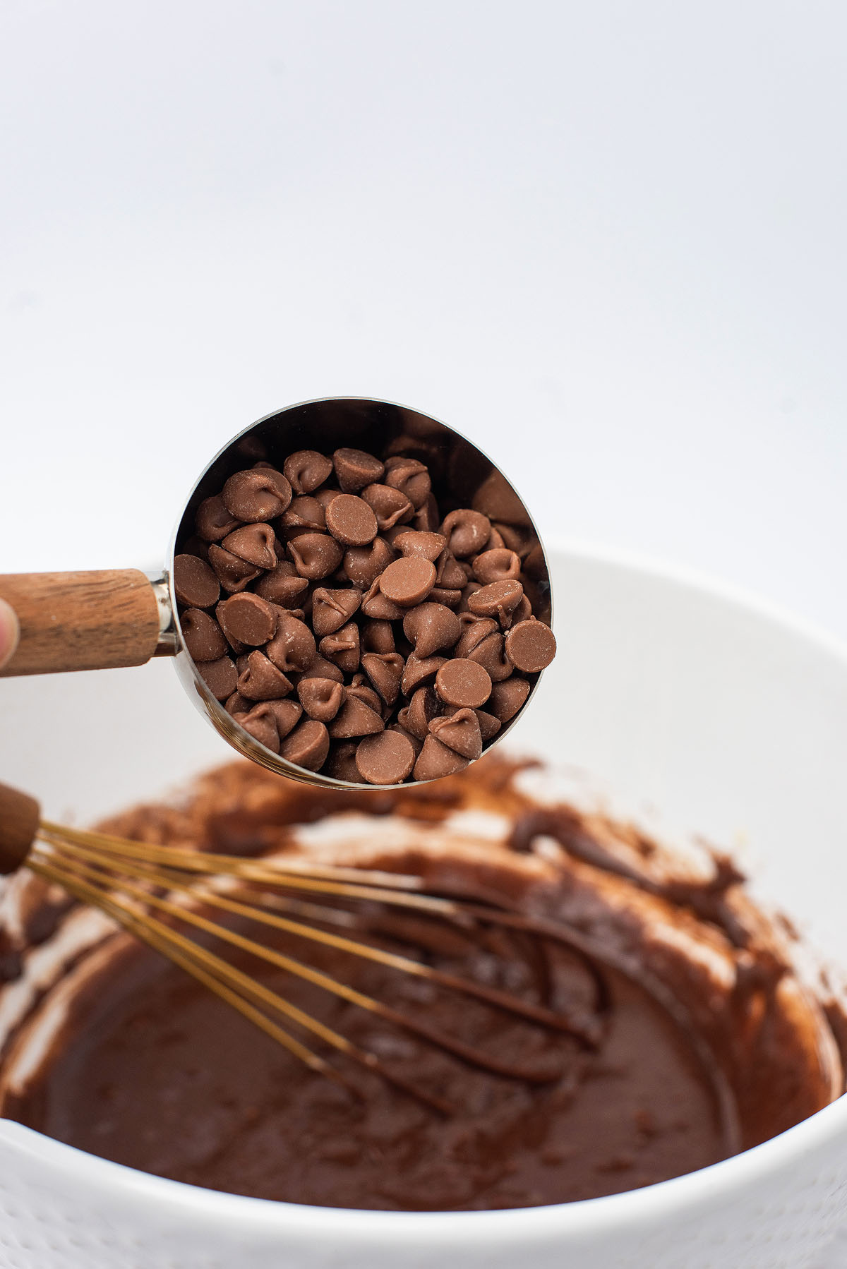 A measuring cup of chocolate chips held over a bowl of chocolate batter with a whisk inside—perfect for making Valentine brownies.