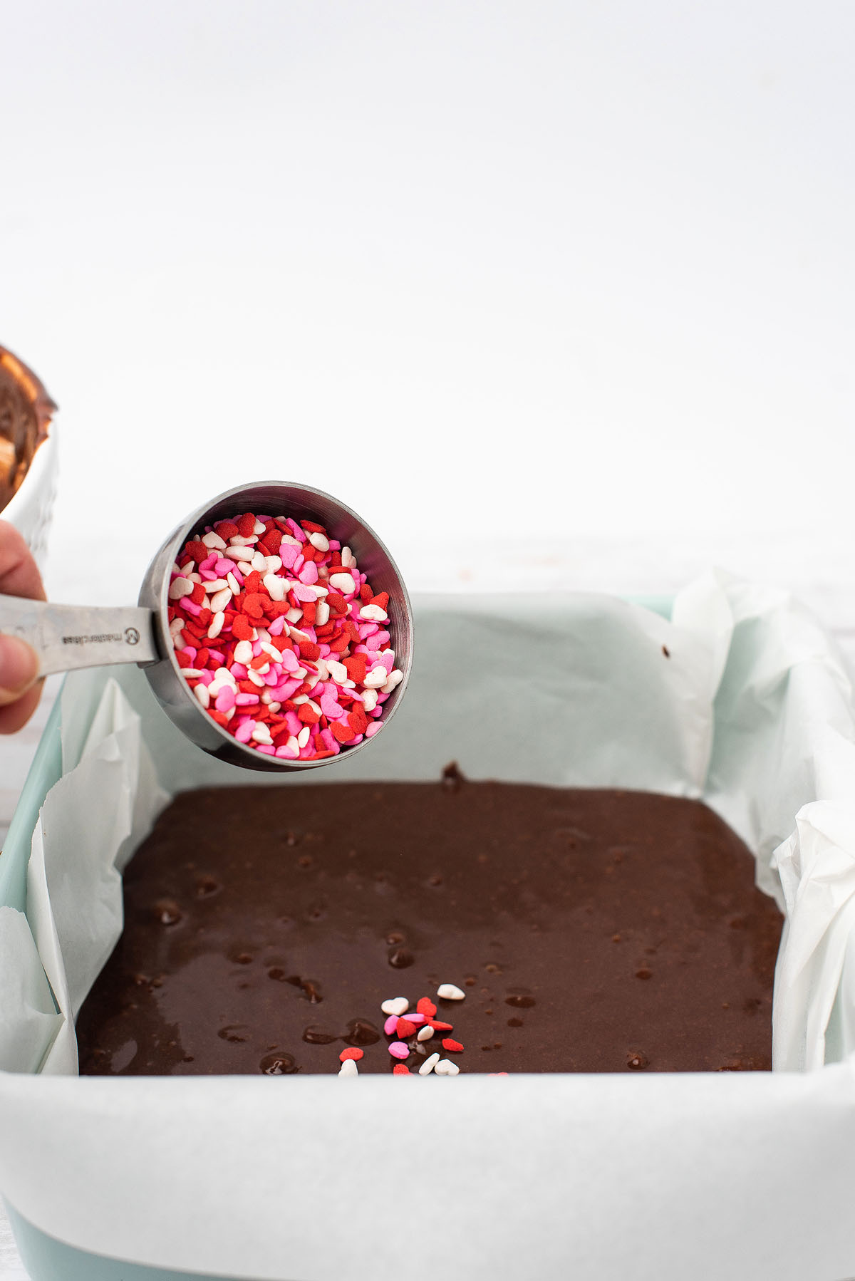 A hand holding heart-shaped sprinkles hovers over a pan of chocolate batter lined with parchment paper, ready to create festive Valentine brownies.