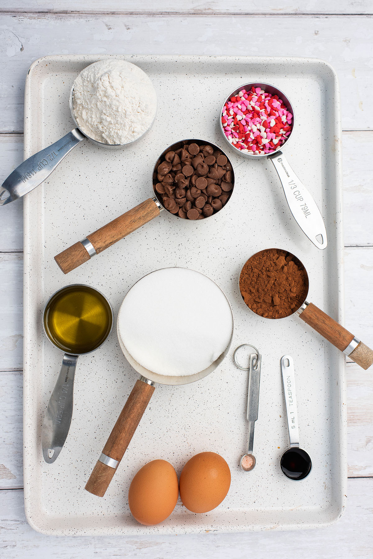 Baking ingredients on a tray—flour, sugar, cocoa, chocolate chips, sprinkles, eggs, oil, vanilla, and salt—are ready to create the perfect Valentine Brownies for a sweet Valentine treat.