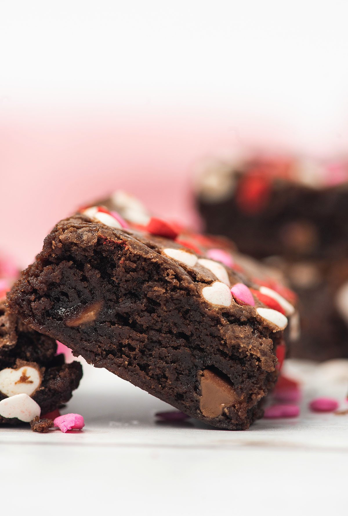 A chocolate brownie topped with pink, red, and white heart-shaped sprinkles makes the perfect Sweet Valentine treat on a white surface.
