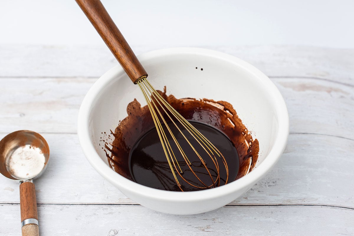 A whisk in a white bowl with chocolate batter sits on a light wooden surface beside a measuring spoon, ready to make delicious Valentine brownies.