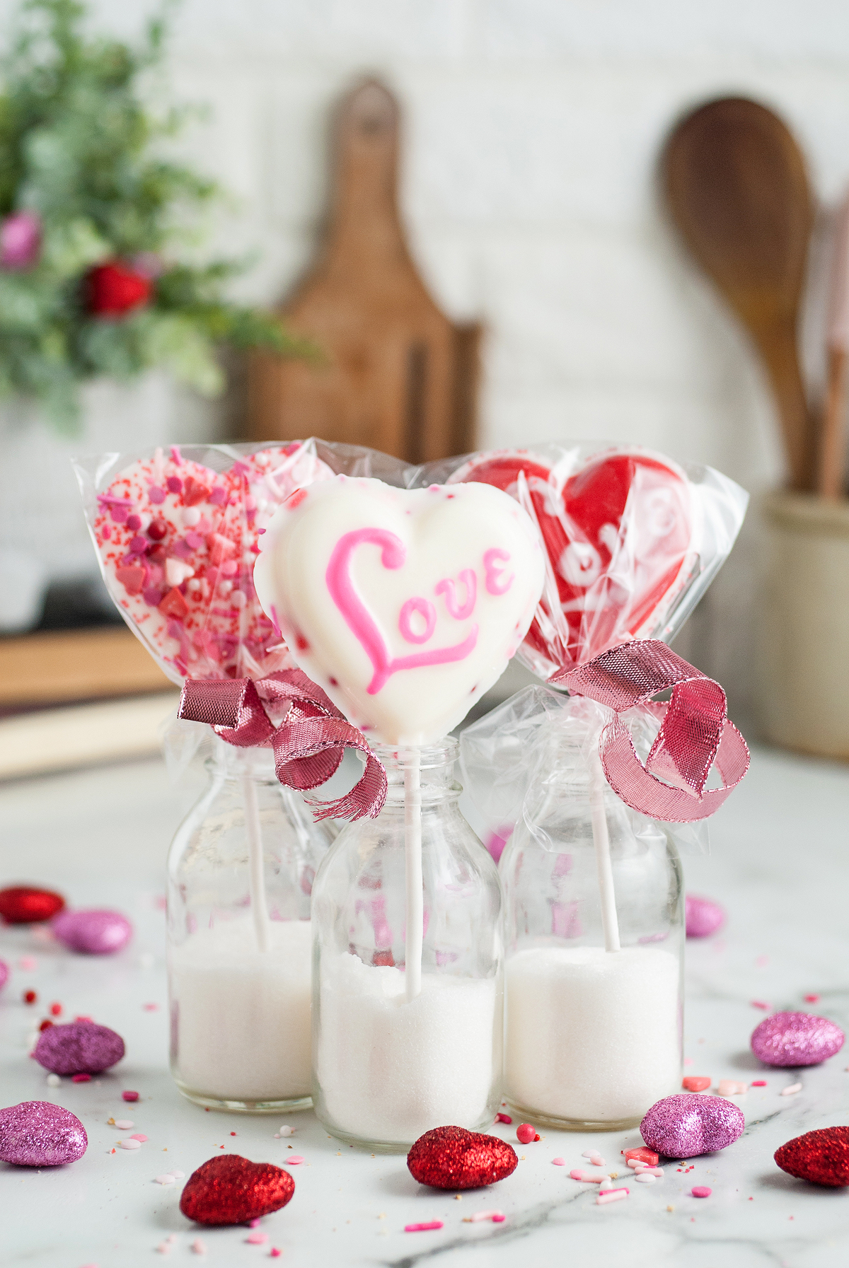 Three heart-shaped decorated lollipops in small glass bottles with pink ribbons, surrounded by glitter hearts—perfect Valentine Lollipop Treats for gifting or displaying your favorite chocolate heart lollipops.