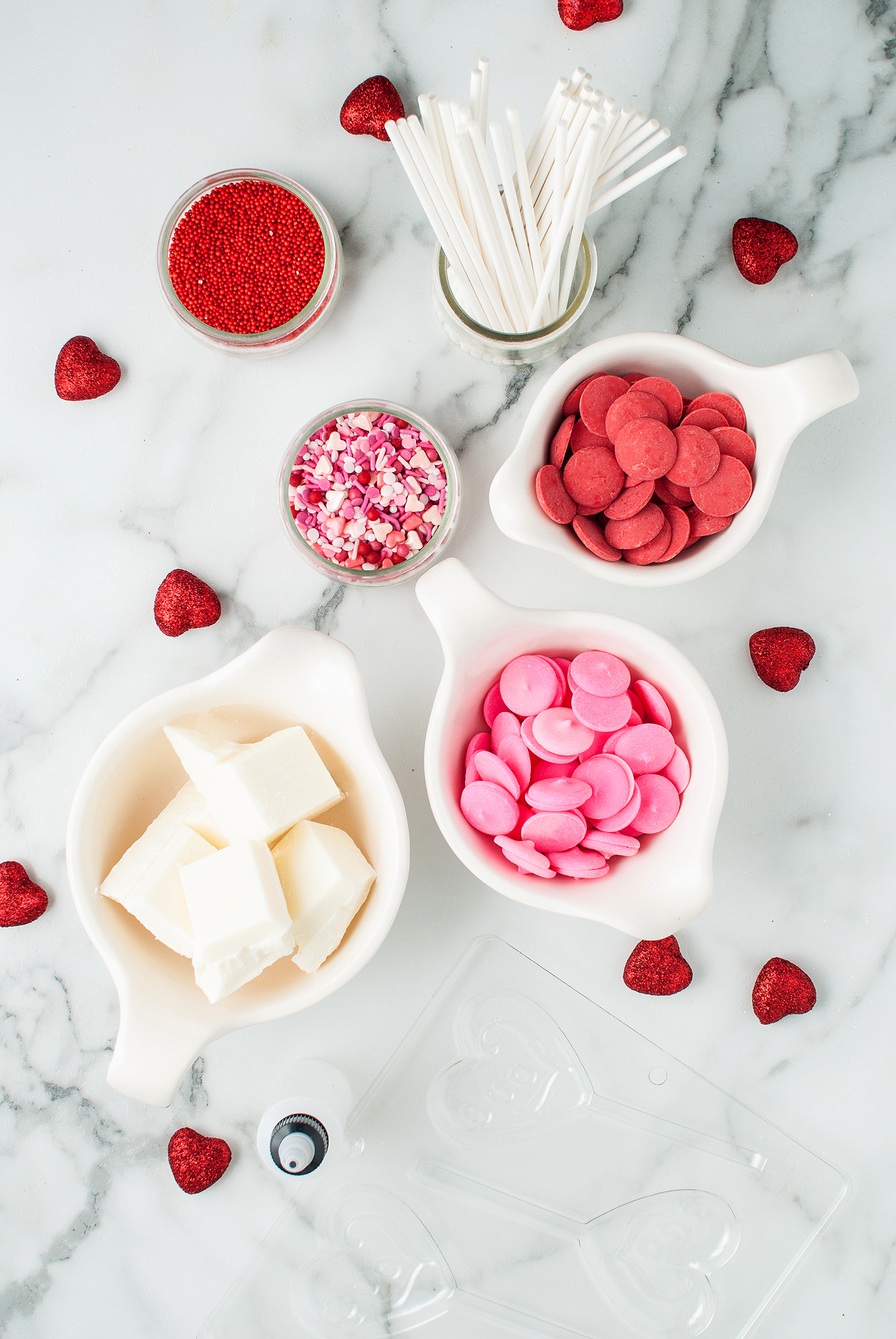 White bowls of white chocolate, pink and red candy melts, sprinkles, and cake pop sticks on a marble surface—perfect supplies to make chocolate lollipops with molds or create festive chocolate heart lollipops.