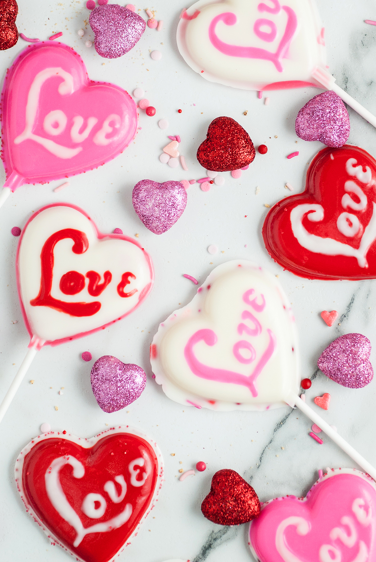 Heart-shaped Valentine Lollipop Treats with the word "Love" and glittery hearts rest on a marble surface—perfect inspiration if you want to make chocolate lollipops with molds at home.