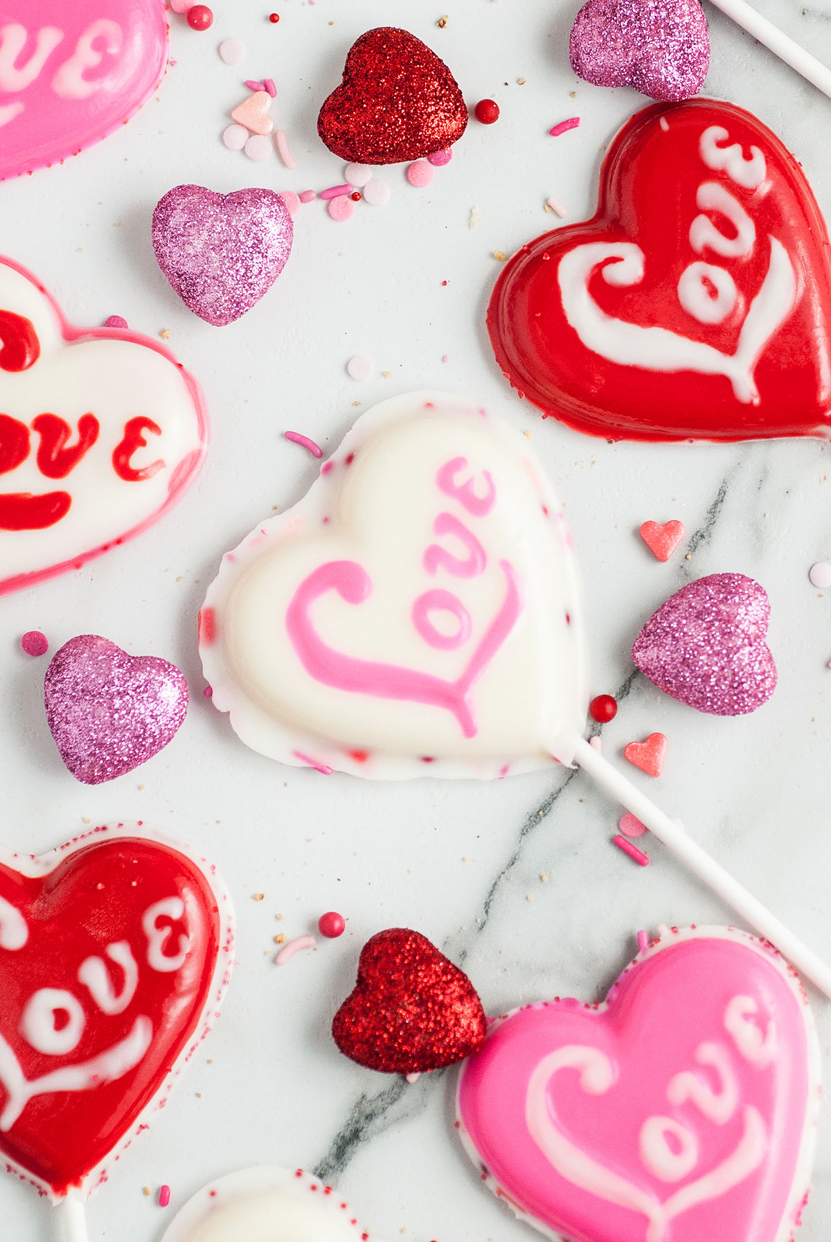 Heart-shaped lollipops with "love" written on them, surrounded by glittery heart decorations on a white surface—perfect Valentine Lollipop Treats for celebrating sweet moments.