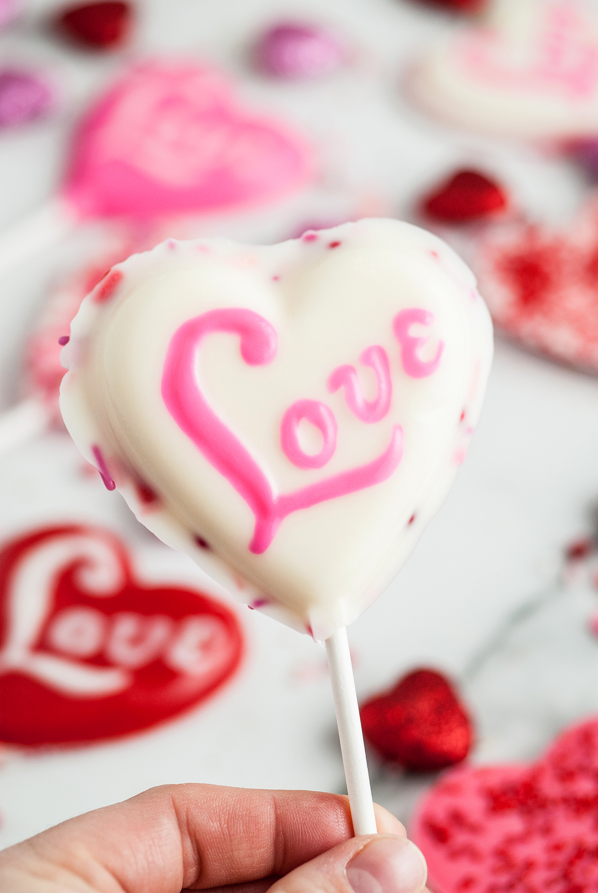 A hand holds a white heart-shaped lollipop with "Love" written in pink icing, perfect as a Valentine’s Day treat or inspiration to make chocolate lollipops with molds for loved ones.