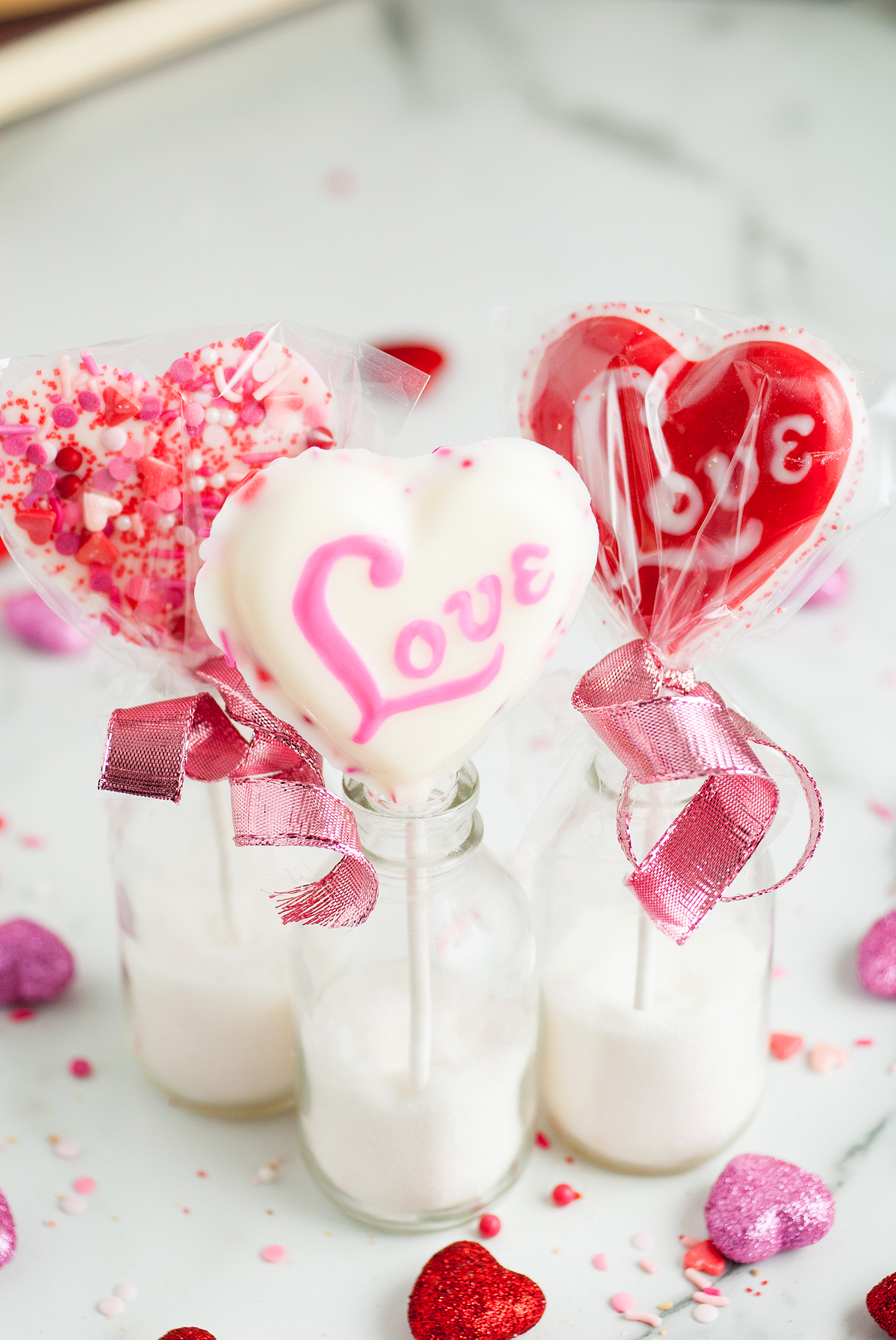Three heart-shaped Valentine Lollipop Treats with "Love" written on them, nestled in bottles with pink bows and sprinkles—a sweet Valentine’s Day treat sure to delight.