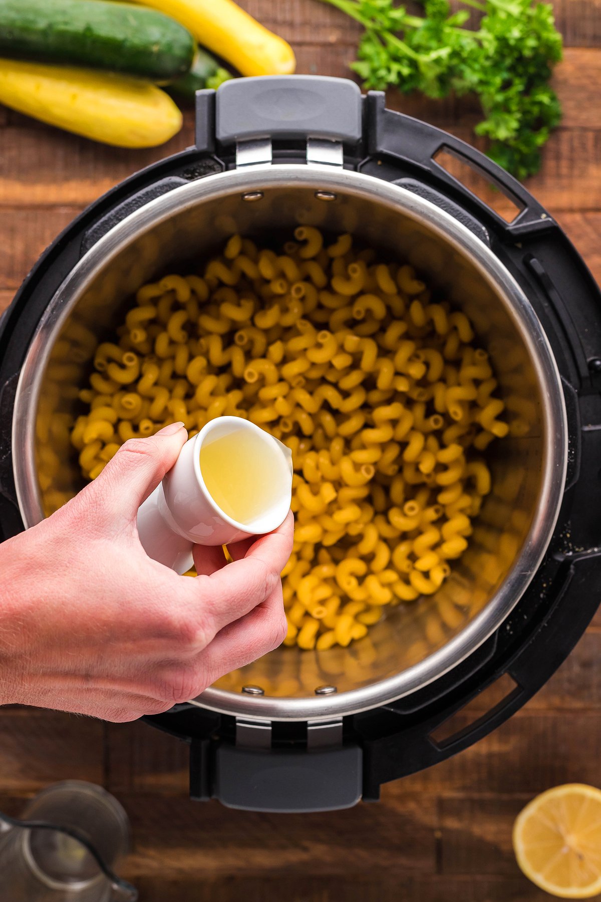 A hand pours liquid from a small cup into an Instant Pot filled with dry elbow macaroni, preparing an easy instant pot pasta dinner.