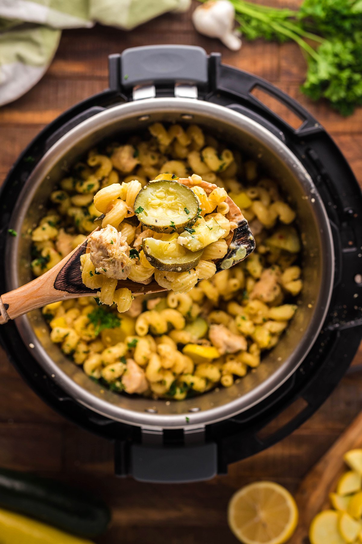 A wooden spoon holds cheesy pasta with chicken and vegetables, featuring zucchini, over a pot on a wooden table.