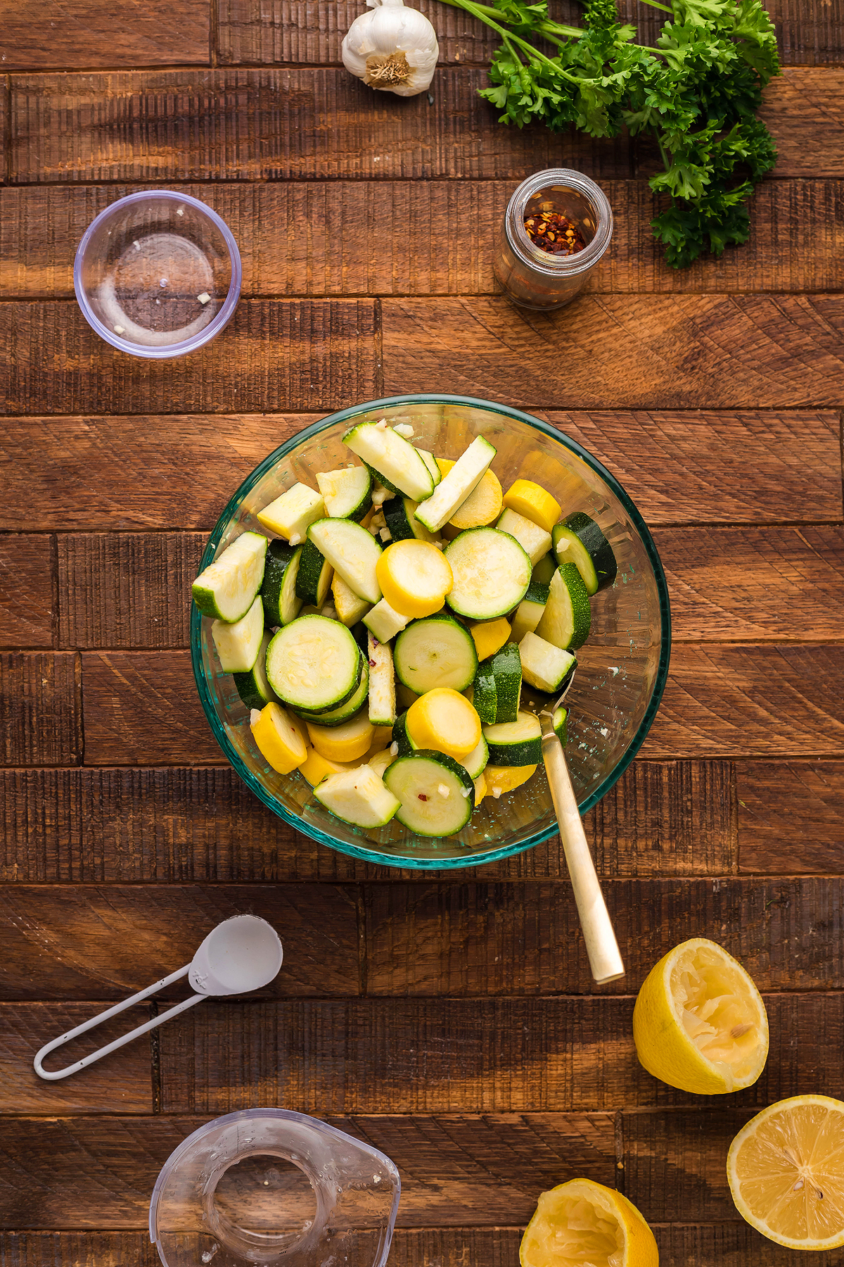 Glass bowl with sliced zucchini and squash, perfect for preparing a fresh Pasta with Zucchini and Squash, surrounded by parsley, garlic, seasonings, and lemon on a wooden table.