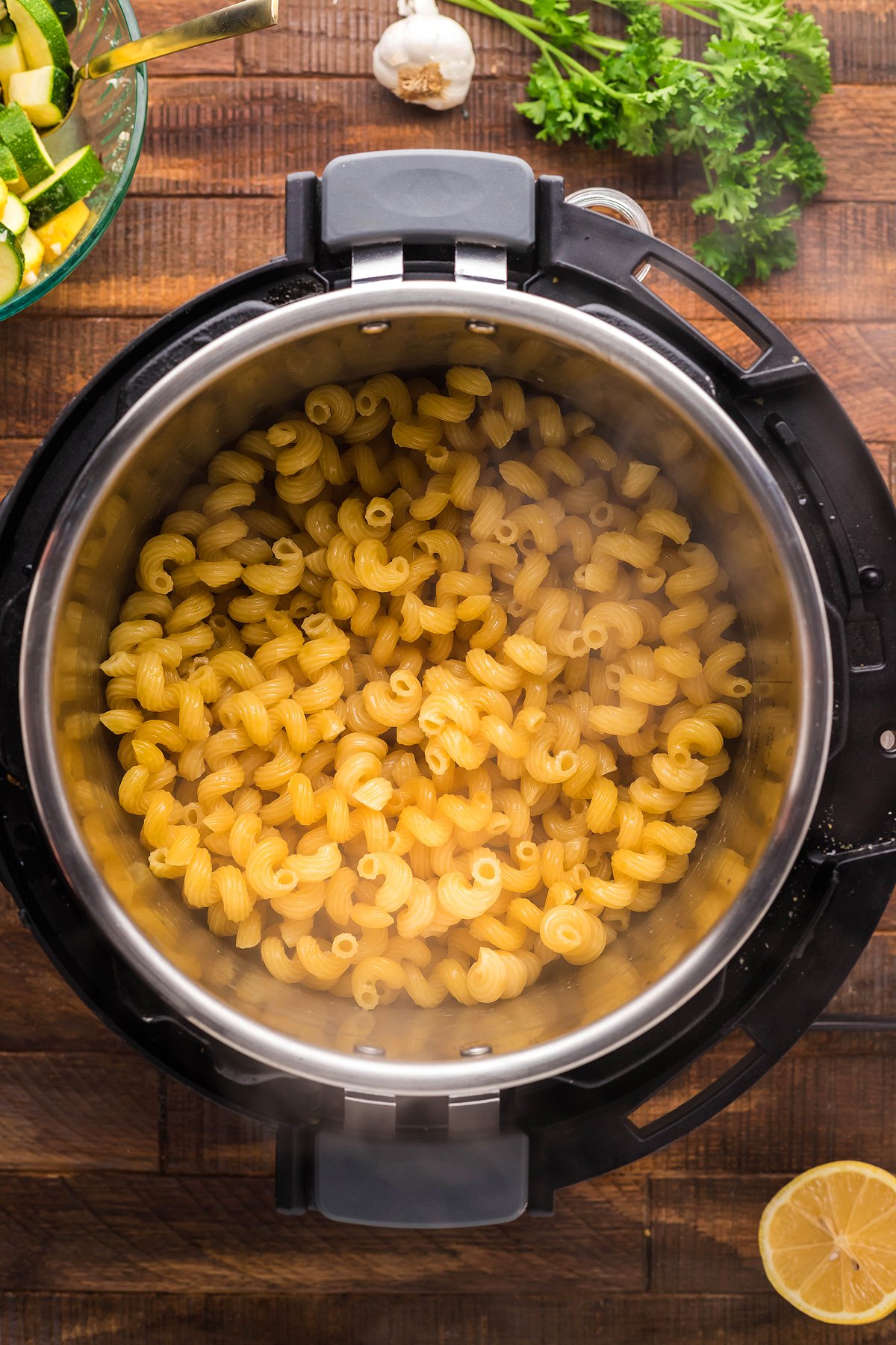 Cavatappi pasta cooking in an Instant Pot for a delicious instant pot pasta dinner, surrounded by lemon, parsley, garlic, and vegetables on a wooden table.