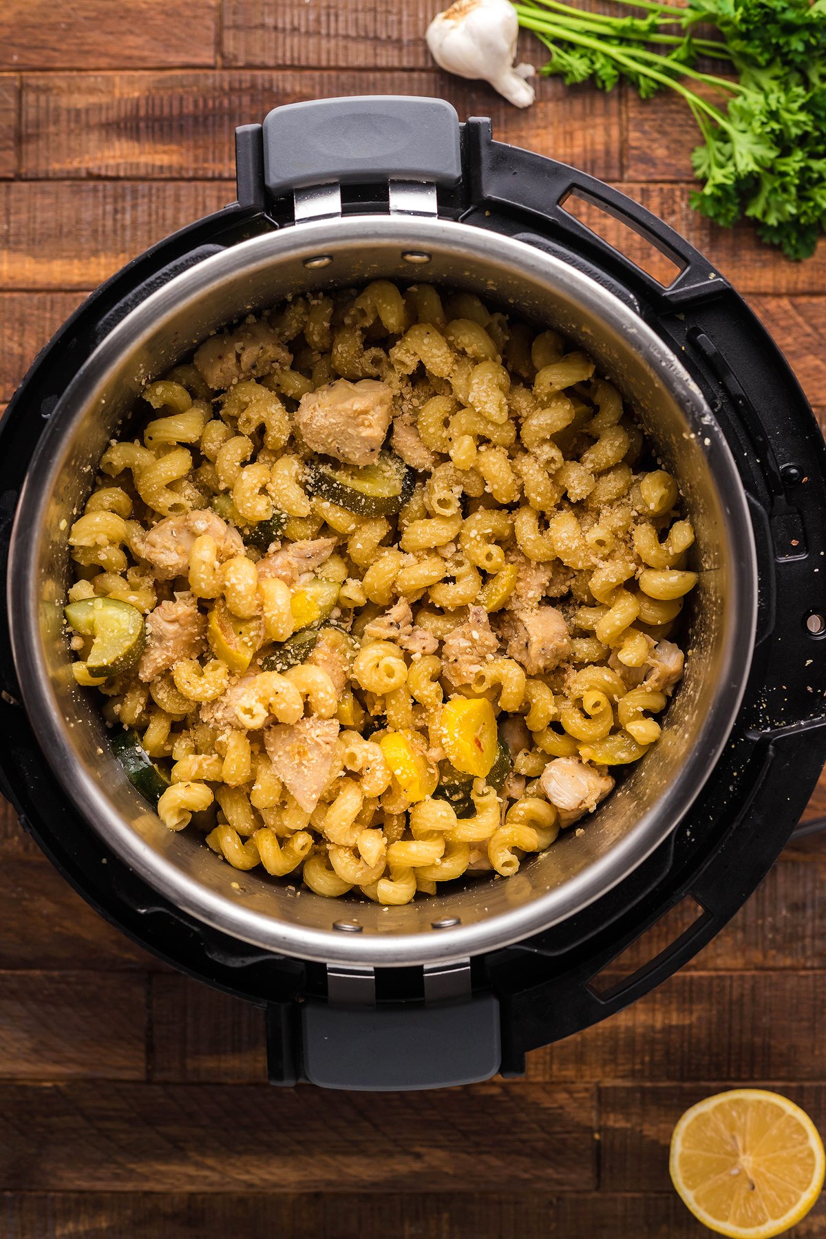 Instant Pot Lemon Chicken Pasta features cooked pasta with chicken and vegetables, served on a wooden surface alongside fresh herbs, garlic, and lemon for a bright instant pot pasta dinner.