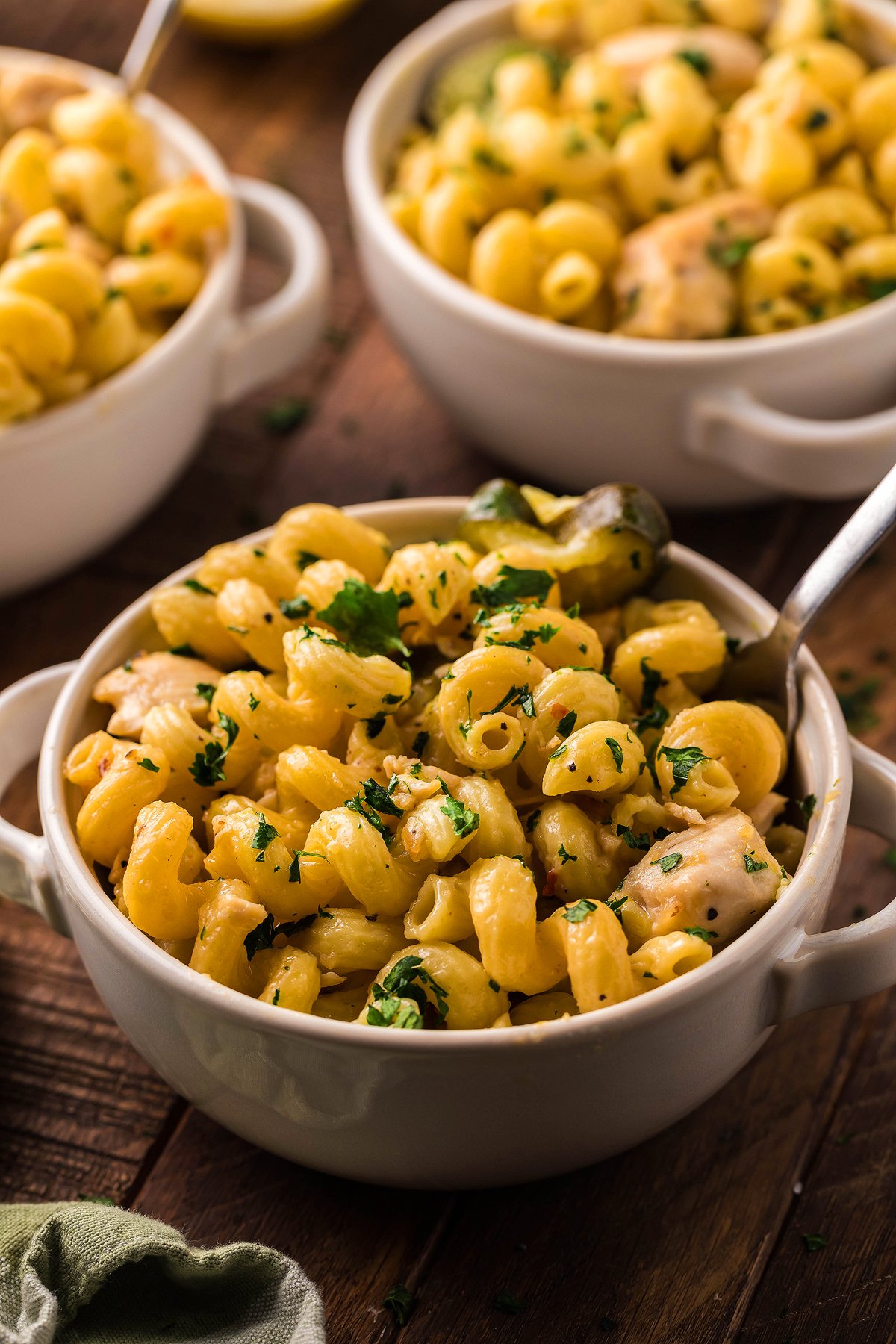 Three bowls of creamy pasta with chicken and vegetables, garnished with fresh herbs, are served on a wooden table with spoons.