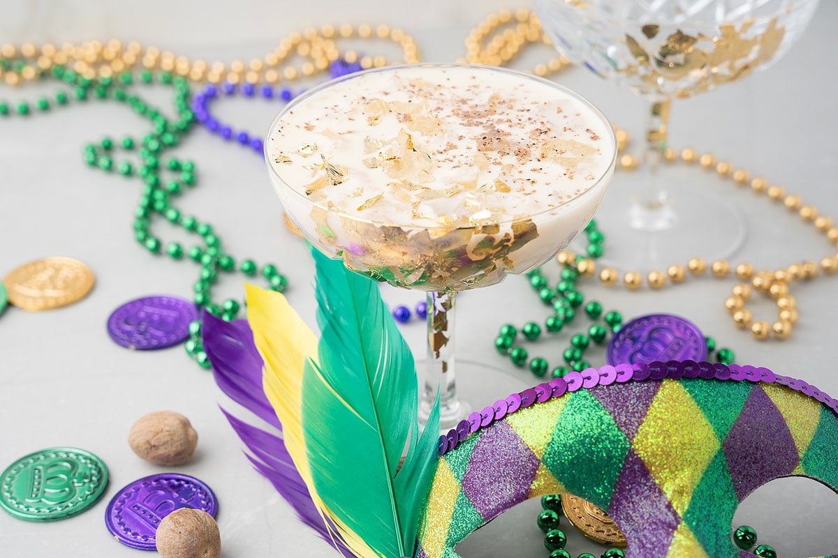 A festive Mardi Gras cocktail adorned with gold flakes, Mardi Gras beads, coins, feathers, and a decorative mask sets the perfect scene for any Mardi Gras party.