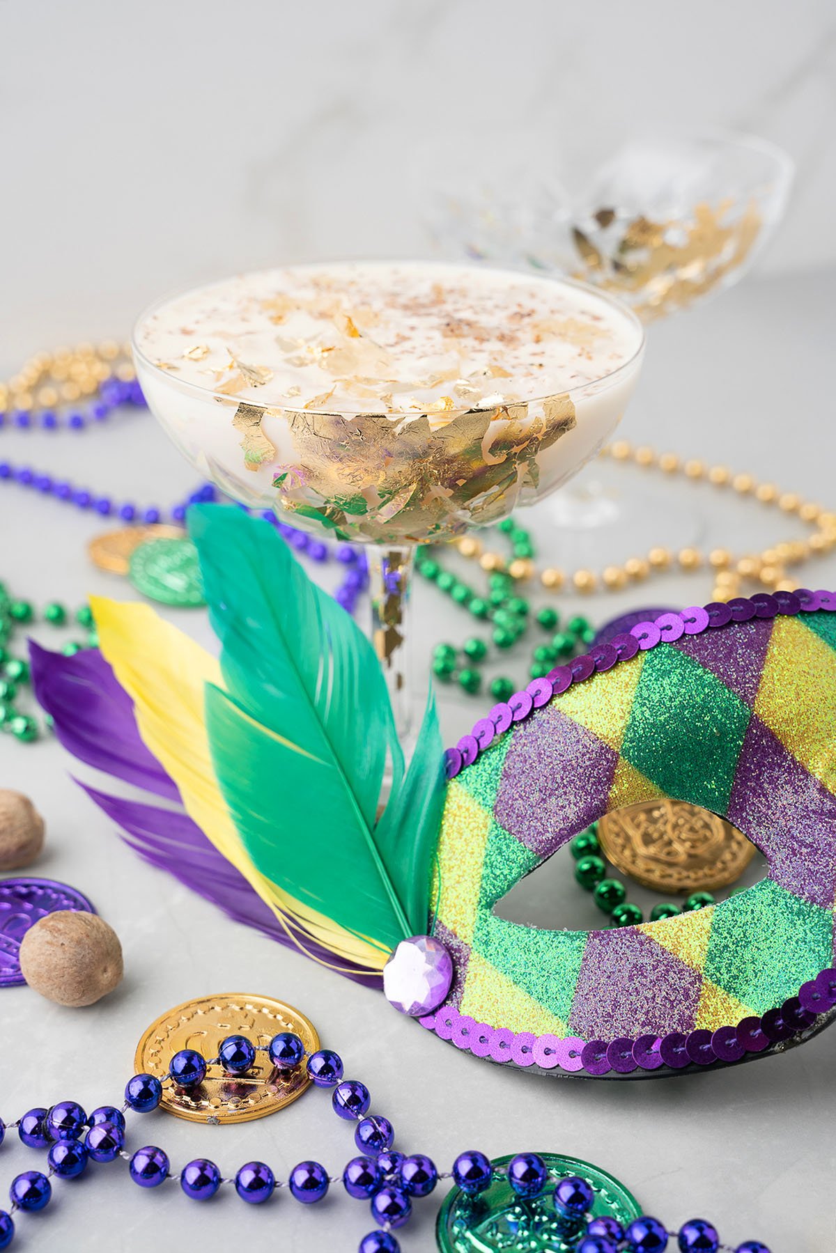 A festive Mardi Gras cocktail adorned with gold flakes, beads, coins, a feather, and a glittery mask—perfect for celebrating New Orleans style with a delicious Brandy Milk Punch twist.