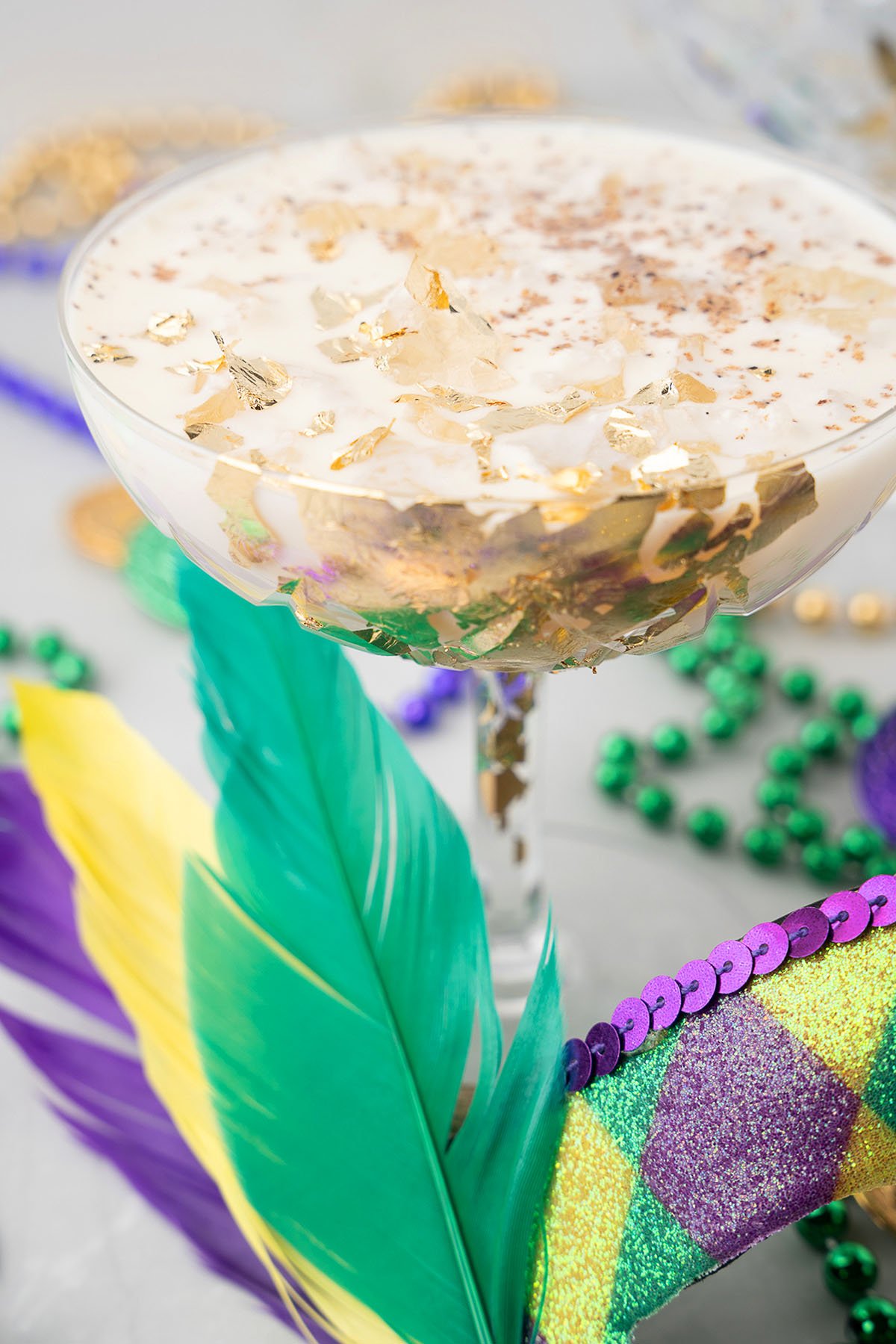 Festive Mardi Gras cocktail with gold flakes, surrounded by Mardi Gras beads, feathers, and a colorful mask—perfect for any lively Mardi Gras party.