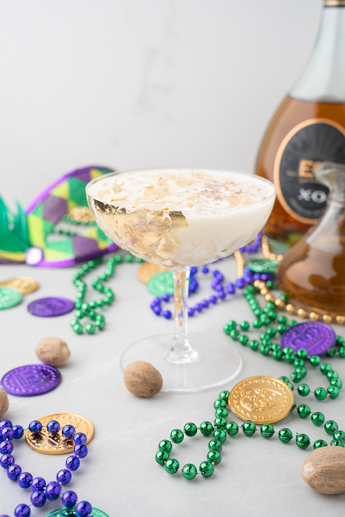 A creamy New Orleans milk punch in a coupe glass, surrounded by Mardi Gras beads, coins, and a liqueur bottle—perfect for your next Mardi Gras party.