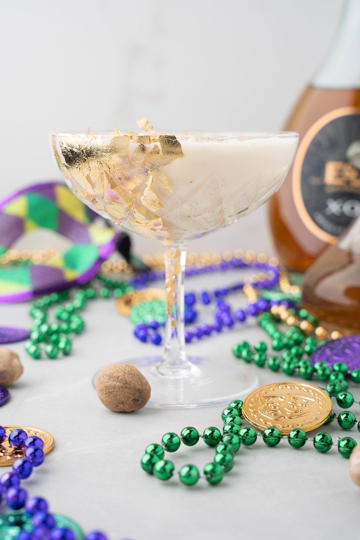 A festive Mardi Gras cocktail in a coupe glass with gold leaf, surrounded by Mardi Gras beads and coins, featuring homemade vanilla bean simple syrup for an extra touch of indulgence.