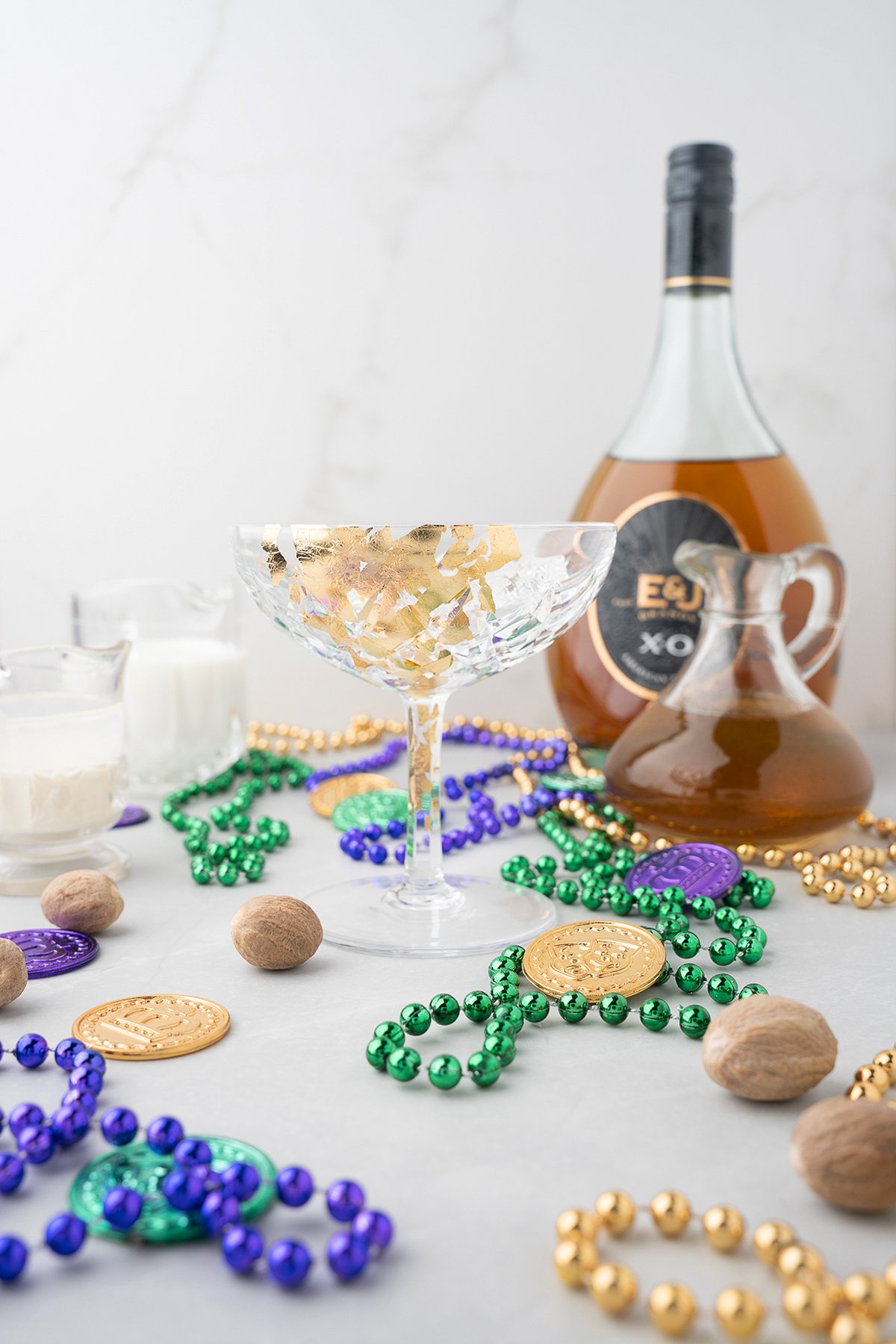 A festive drink setup perfect for a Mardi Gras party, featuring beads, gold coins, a cocktail glass, and all the fixings for classic Brandy Milk Punch—brandy, cream, and nutmeg.