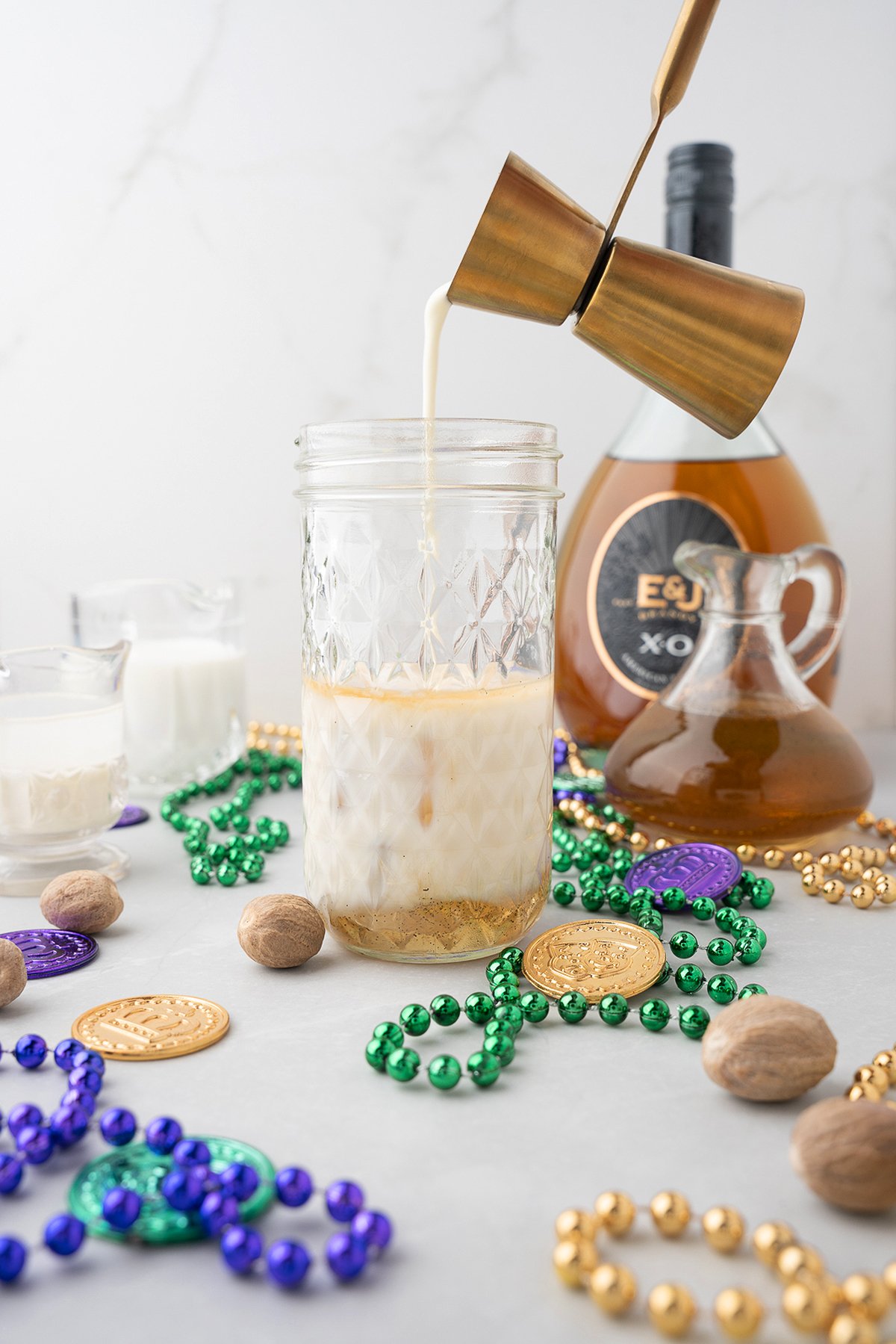 A creamy Brandy Milk Punch is poured into a glass, surrounded by Mardi Gras beads, gold coins, and a brandy bottle—perfect for a festive Mardi Gras party.