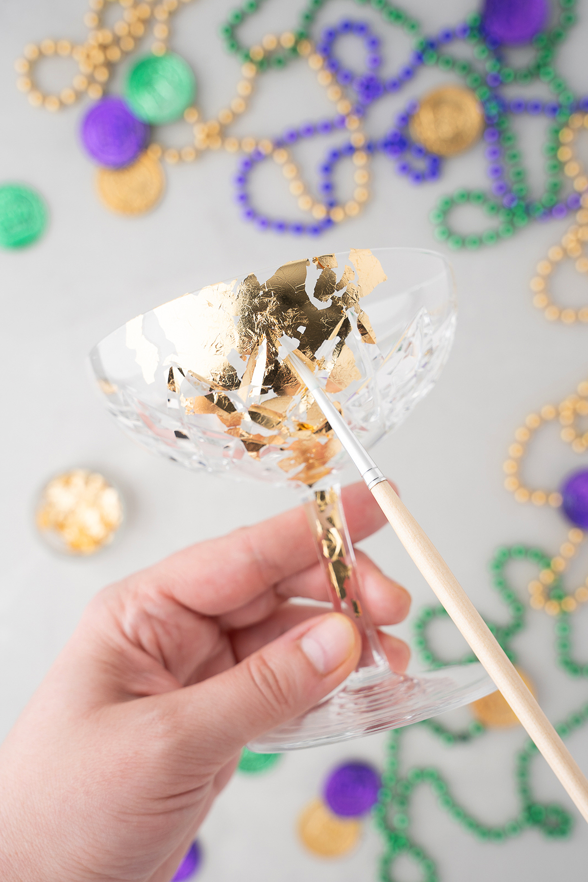 A hand applies gold leaf to a glass with a brush; Mardi Gras beads are scattered in the background, highlighting the festive spirit of a New Orleans milk punch at a Mardi Gras party.