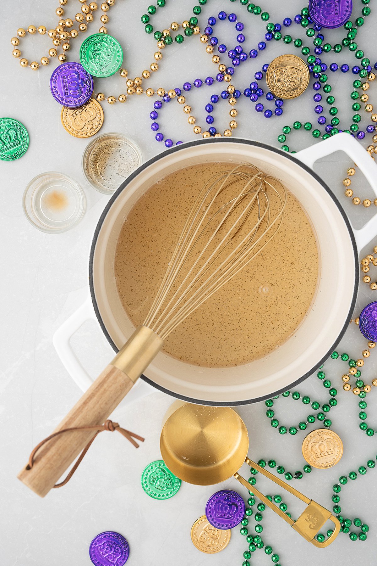 A pot of brown Brandy Milk Punch being whisked, surrounded by Mardi Gras beads and colorful coins on a gray surface—perfect for a festive Mardi Gras party.