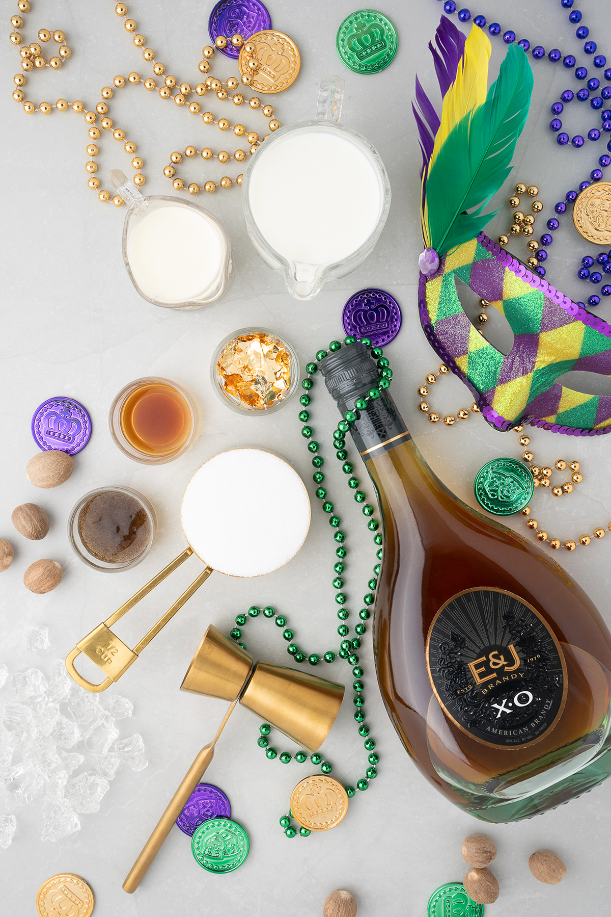 Flat lay of Mardi Gras beads, coins, mask, a bottle of E&J XO, milk, homemade vanilla bean simple syrup, and other cocktail ingredients on a gray surface—perfect for mixing up a classic New Orleans milk punch.