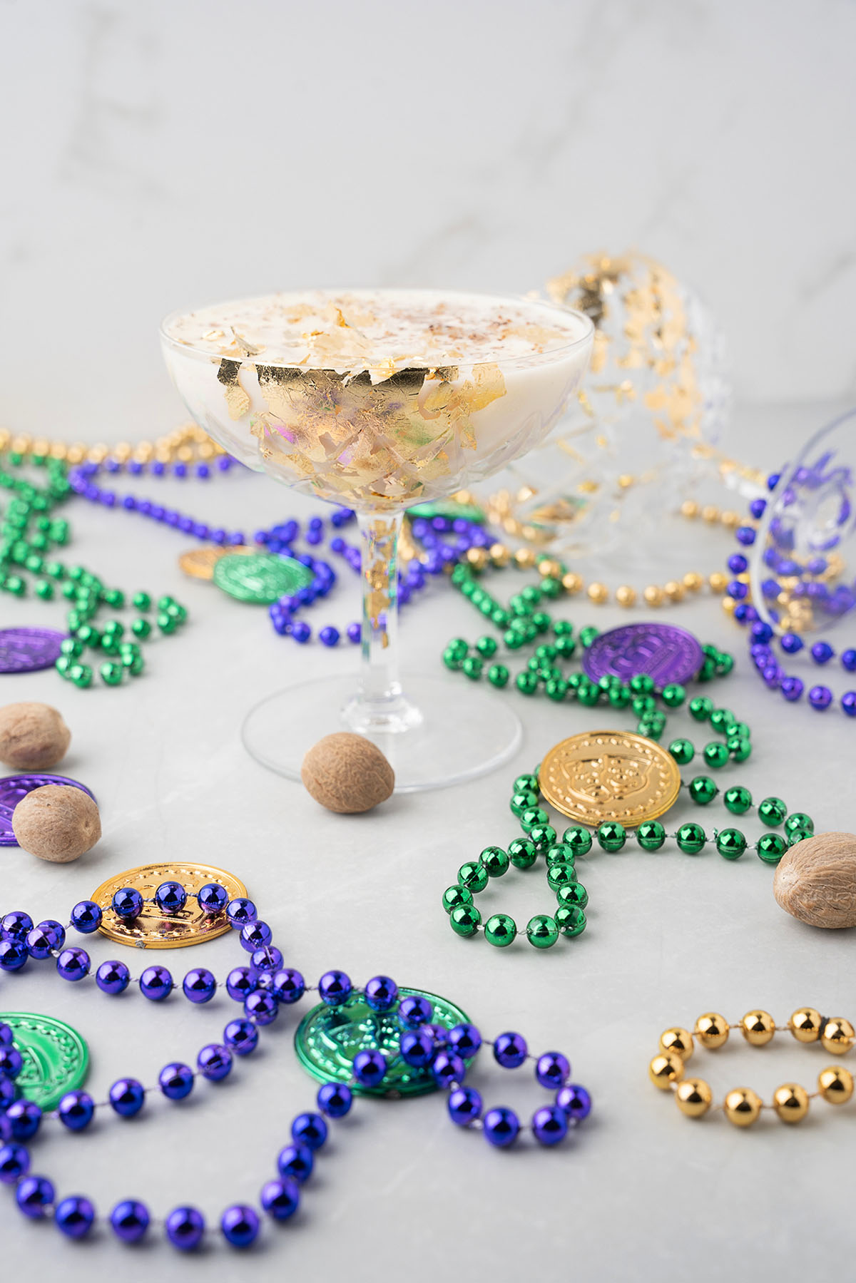 A glass of dessert, inspired by New Orleans Milk Punch, is surrounded by Mardi Gras beads, gold coins, and whole nutmegs on a light surface.